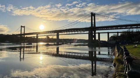 The Tamar Bridge as the sun gets low in the sky and is reflected in the water below the bridge. There is an iron railing in the foreground in front of a grassy area with a bench in it.