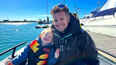 Photograph of Andy Johnson and his son, Oaklen. The pair wear life saving jackets and sit inside a paddle boat. They both wear hooded jackets under their life vests and smile at the camera.