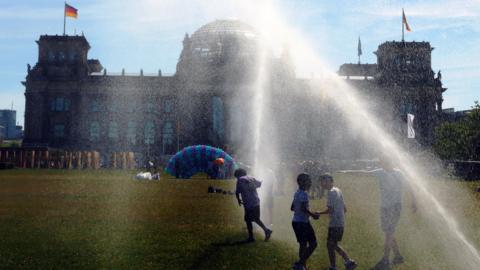 People pass by a watering system in front of the Reichstag on a hot summer day on June 24, 2026 