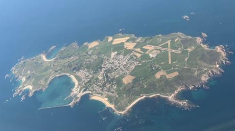 An aerial view of the entirety of Alderney showing the island surrounded by sea. Fields and areas of housing are visible. 