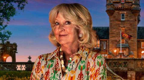 Handout photo of Fiona, one of the contestants in series four of BBC1's The Traitors. Fiona has short blown curled hair, blue eyes and smiles at the camera. She wears a yellow, green and red patterned blouse, a thick shell necklace and diamond earrings. A UGC image of the castle is seen in the background.