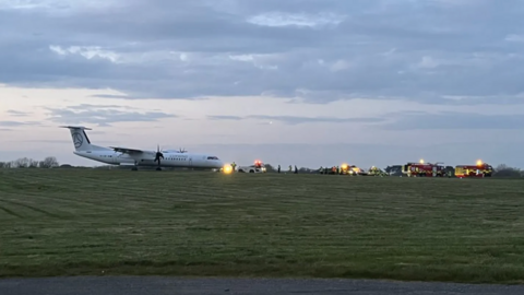 An aeroplane in the distance with emergency vehicles in front of it. Grey clouded skies at sunset behind with grass seen in front.