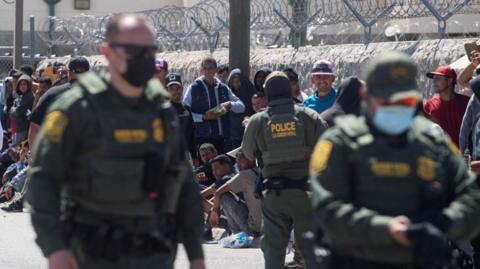 Police officers stand while immigrants wait to be processed by U.S. Customs and Border Protection (CPB) near the border with Mexico