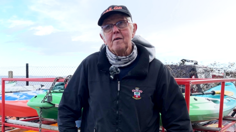 Ian Cobb is on the right of the frame, wearing a cap and a pair of glasses. He is wearing a scarf, a Southampton Football Club rain jacket and blue jeans. Behind him are rows of kayaks and canoes in different colours, and in the background is the Bristol channel.