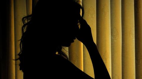 A woman with her head in hands. Only her silhouette is seen in a dark room with yellow curtains.