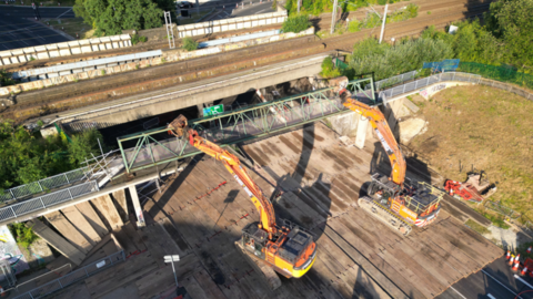 Two cranes are lifted above a bridge. Concrete pillars lay on the ground below along a grass verge.