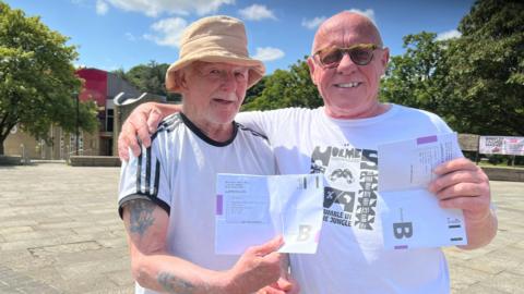 Two middle aged white men in white T shirts holding up envelopes. They stand in what looks like a market square. It's a sunny day and one of them wears a sun hat.