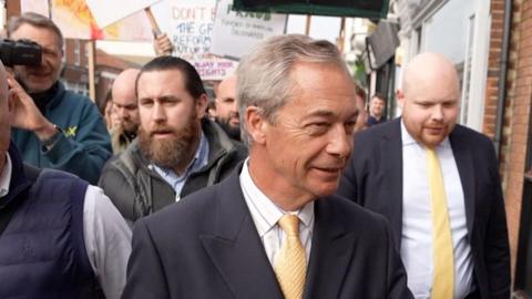 Nigel Farage is wearing a dark blue suit and a yellow tie. He is surrounded by people, one holding a camera. In the background, protest signs are visible.