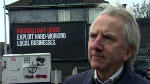 Máirtín Ó Muilleoir with a paramilitary sign in the background. He has white hair, wearing a blue jacket.