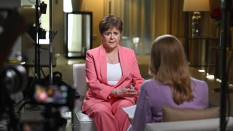 Nicola Sturgeon with her head tilted and a pensive look on her face. She is wearing a pink jacket and has a chain around her neck.