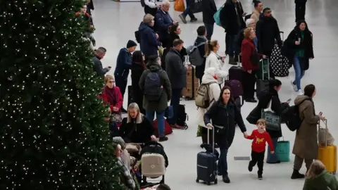 People gather in train station with suitcases, a Christmas tree with lights on it is in the foreground