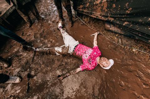 Enter Shikari wearing a pink t-shirt and cream pants lies in a pool of mud while holding his mic in one hand