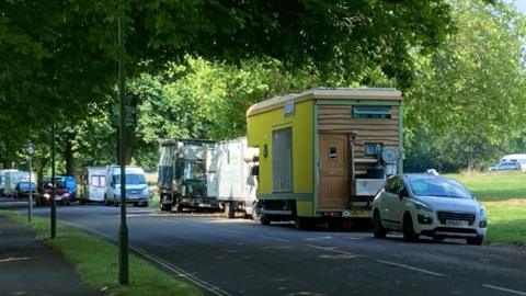 A row of campervans, trucks and cars line a street on the Bristol Downs with a green field in the background.