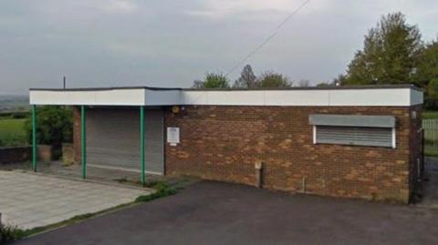A street view of a brick, one-storey building with a white roof that overhangs the front door area, which has a shut roller-blind, and is held up by green poles. There are parking spaces in front of the building and trees and a field beyond.