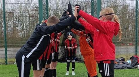 Members of a junior football club wearing red and black coloured football kit. The young football players have formed a human tunnel by joining their hands together and one of their team mates is stood at the entrance of the tunnel.