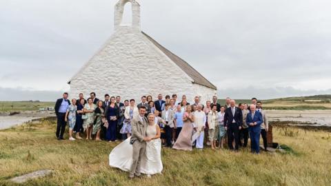 This image shows a couple after getting married, they are stood outside the little church with their wedding guests stood behind them. 