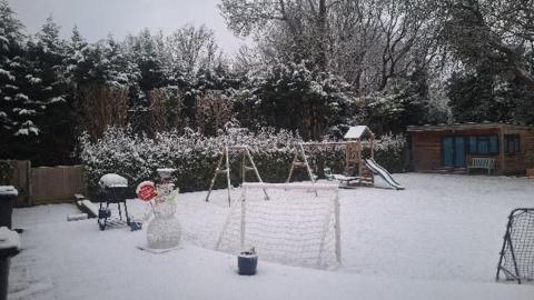 A "stop here santa" Christmas decoration covered in snow looks like a snowman in a garden. There is play equipment and everything is snowy. 