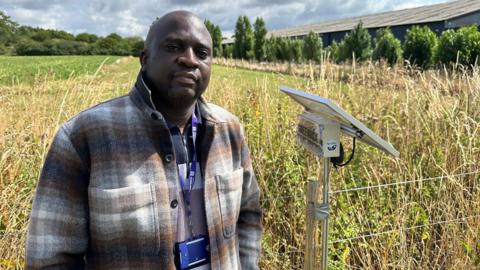 A man standing in a field in front of a wire fence. He is bald and is wearing a white, brown and blue jacket with a purple lanyard around his neck. Next to him is a solar panel on a metal pole which is attached to the fence. It has a box attached to its underside that reads "green box"