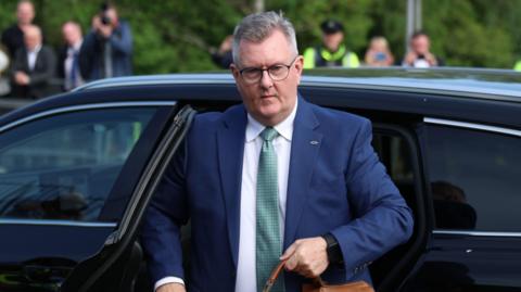 Jeffrey Donaldson, with short grey hair, wearing a navy suit, white shirt, green tie and glasses. He is getting out of a black car. 