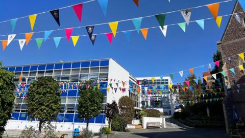Beaulieu Convent School on a bright blue sky day. There are two buildings which are electric blue and feature many windows. There is multi-coloured bunting strung up high above trees and a bench.