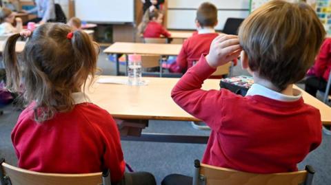 Pupils in a primary school in Glasgow. Two children - a girl and a boy -have their backs to the camera. They are both wearing a red jumper and white polo shirt. They are sitting in small wooden chairs at a large desk. There is classroom activity in the front of them.
