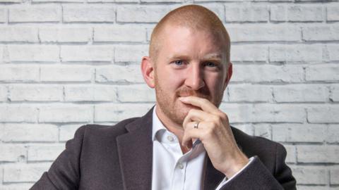 A man with very close-cropped red hair and reddish stubble is in front of a white brick wall. He is wearing an open-necked white shirt and a dark blazer. He is resting his chin on his hand with a finger in front of his mouth.