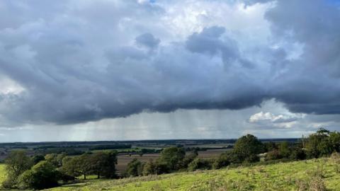 A huge blue, grey cloud fills the sky over green fields and trees. Rain can be seen falling near the horizon.