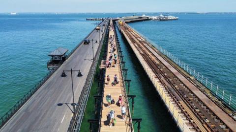 A pier over the sea. It is divided into three seperate sections. There are people walking up the middle section, and the section on the right has railway tracks on it.