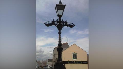 An ornate black lamp post with a crossbar with foliage decoration and the lamp topping the post. Several low storey buildings are in the background.