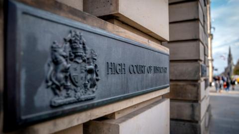 A dark grey metal sign on the sandstone block wall of the High Court in Edinburgh. The sign has a royal crest and the words: "High Court of Justiciary".