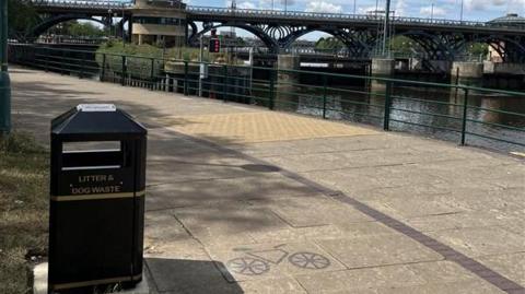 A new black bin is in the bottom right with 'Litter & Dog Waste' written on the side. It is next to a cycle path and footpath with a river on the right side.