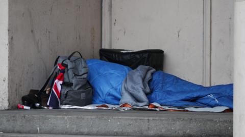 A blue tent depciting a rough sleeper with a grey rucksack nearby with part of a Union Jack flag showing attatched.