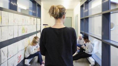 A blonde female teacher, wearing a black top, with her back to the camera facing four school children sitting in a school corridor. They all appear to be looking at phones.