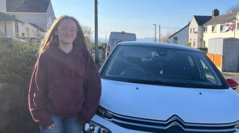 Tilly Kay stands next to her car. She has long brown hair and wears glasses. She is wearing a burgundy hoodie and denim jeans. Her car is white and is parked on the street in a housing estate.
