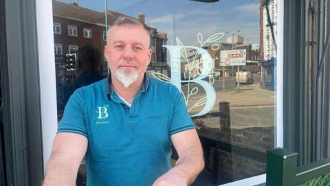 A man with grey hair and a white beard stands in front of a large glass window wearing a blue polo top.
