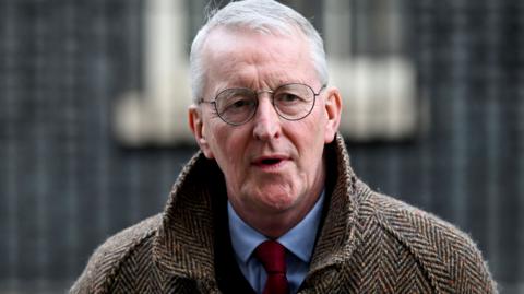Northern Ireland Secretary Hilary Benn , he has short white hair, round-rimmed glasses and is wearing a brown herringbone-patterned overcoat over a blue shirt and red tie