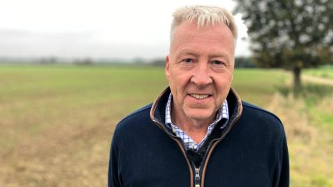 James Alston, a farmer, standing in a field smiling at the camera in a blue square check shirt and a blue zip-up top.