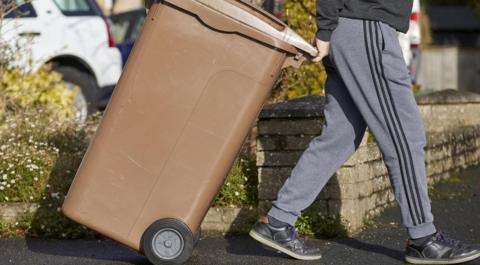 A person wearing a dark grey sweater, grey joggers with three black vertical stripes on, and black trainers wheeling a brown bin on the pavement outside. 