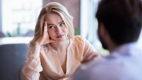 a young woman sits across from a man at a table. Her head is resting on her hand. She looks annoyed