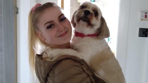 Blonde girl with high ponytail looking at the camera. She is wearing a green puffer jacket and holding a small white and tan dog. The dog is wearing a red collar.