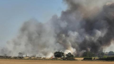 View across agricultural fields with flames and a large column of smoke visible in the distance