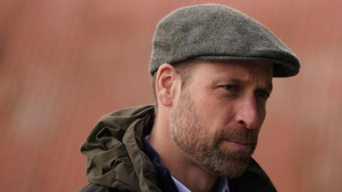 The Prince of Wales, bearded and wearing a grey flat cap and a hooded jacket, looks off-camera.