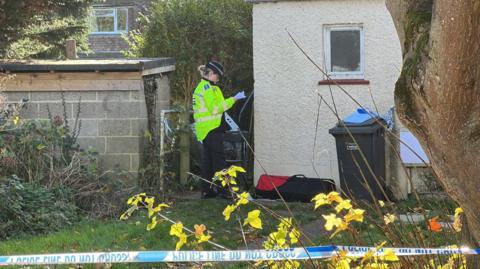 A policewoman in a hat and high-vis jacket looking at paper while standing by a building which is behind blue and white police tape.