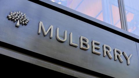 A Mulberry store signage with thin capital letters and a mulberry tree on a dark wood background. Above the sign there is a window, in which a designed handbag can be seen.