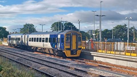 A blue and white, two-carriage Northern train pulled up to the station platform. There are yellow barriers erected on the platform, with a worker wearing a high-vis uniform standing on it too. There are about a dozen poles rising from the station grounds, with CCTV and street lights.