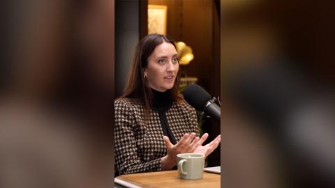 A woman with long, brown hair talks to someone off-camera.
