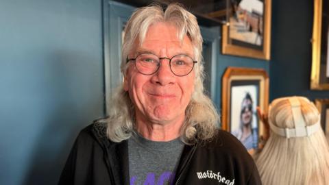 A man with long grey hair cut into a mullet style. He has black rimmed round glasses and is wearing a black Motorhead hoodie with a grey and purple Black Sabbath t-shirt underneath.