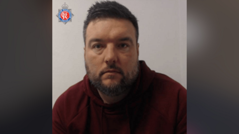 Richard Shaw has a maroon hoodie, brown hair and a brown beard.