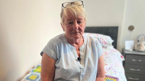 Woman with short blonde hair black glasses on head light blue blouse sat on single bed with blanket on top