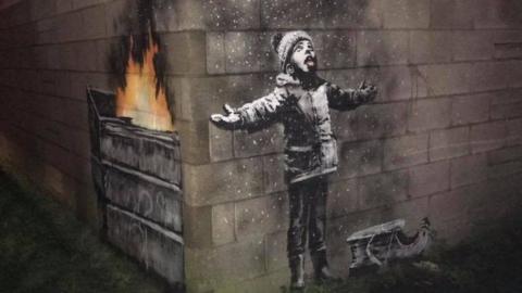 Banksy drawing across two walls at right angles - the right side shows a young child with his arms splayed, a sled at his feet and tongue out looking at what looks like falling snow. The left side reveals the snow is in fact ash from a fire burning in a dump truck.